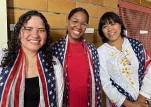 Three adult students who are women stand in a row, in front of a brown tiled wall. All are wearing American flag-printed scarves over their shoulders, and are looking at and smiling for the camera.
