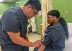 Two adult nursing students, both dressed in dark blue scrubs and wearing eyeglasses, practice the skill of taking a patient's blood pressure. One student is standing and holds a stethoscope to the other seated student's arm, which is wrapped in a blood pressure cuff. The wall behind them is bright green. 