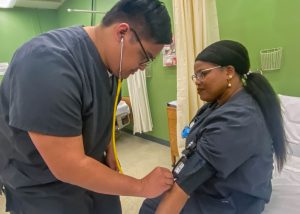 Two adult nursing students, both dressed in dark blue scrubs and wearing eyeglasses, practice the skill of taking a patient's blood pressure. One student is standing and holds a stethoscope to the other seated student's arm, which is wrapped in a blood pressure cuff. The wall behind them is bright green.