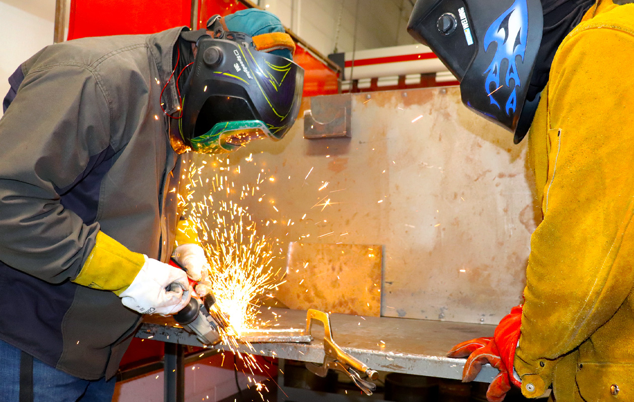 Student welding as teacher looks on, both wearing helmets.