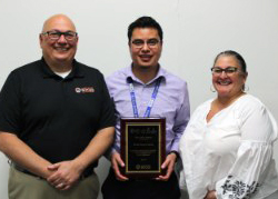 Capital Region BOCES' Senior Executive Officer Joseph Dragone, Network Technician Daniel Solares and District Superintendent Anita Murphy stand together in a row. Daniel is holding a plaque that recognizes his contributions to the organization. All three are looking at and smiling for the camera. 