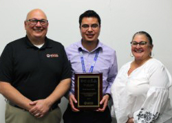 Capital Region BOCES' Senior Executive Officer Joseph Dragone, Network Technician Daniel Solares and District Superintendent Anita Murphy stand together in a row. Daniel is holding a plaque that recognizes his contributions to the organization. All three are looking at and smiling for the camera.