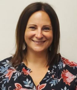 School Counselor Jessica Iorio, who has long straight brown hair and is wearing a floral patterned blouse looks at and smiles for the camera.