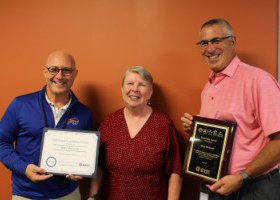 Assistant Superintendent Michael Doughty, Data Warehouse Manager Mary McGeoch, and Program Manager Jeff Luks stand in a row. Mike Doughty is holding a certificate and Jeff Luks is holding a plaque that they presented to Mary in recognition of her Extra Mile Award. All are looking at and smiling for the camera.