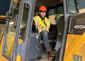 Career and Technical Education Student Alex Stapf, who is wearing a flourescent orange hard hat, black sunglasses, a yellow safety vest and jeans, sits in a large, yellow, construction vehicle in the garage of a Capital Region BOCES construction and heavy equipment classroom. Stapf is turned forward and looks at the camera.