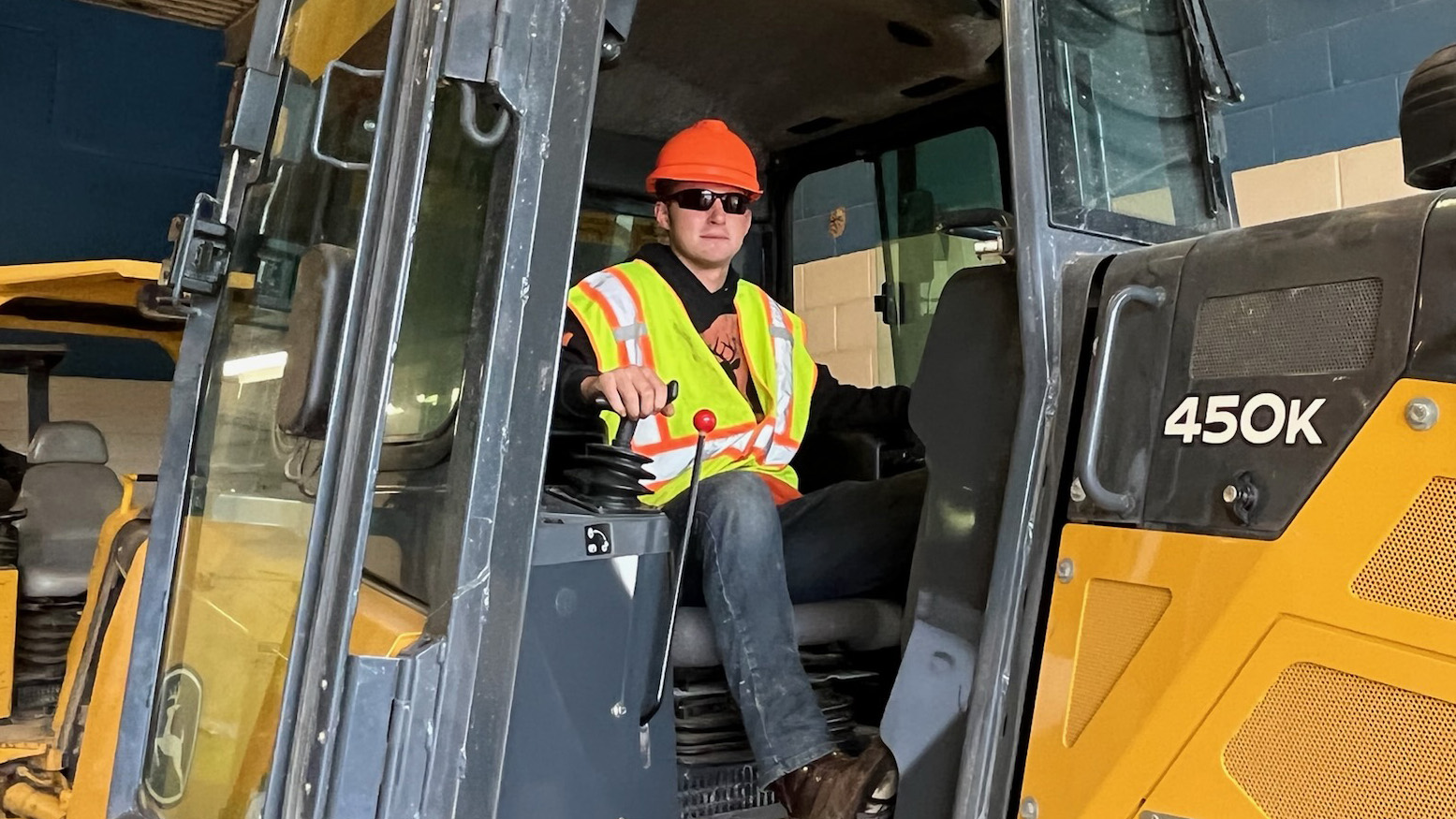 Career and Technical Education Student Alex Stapf, who is wearing a flourescent orange hard hat, black sunglasses, a yellow safety vest and jeans, sits in a large, yellow, construction vehicle in the garage of a Capital Region BOCES construction and heavy equipment classroom. Stapf is turned forward and looks at the camera.
