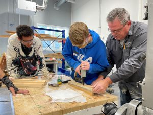 A student in a blue sweatshirt and protective glasses working on wood pieces while another student and instructor look on.
