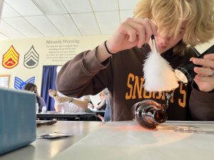 A criminal justice student, wearing a brown sweatshirt, dusts off fingerprints from an amber glass bottle.