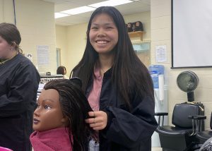 Cosmetology program graduate Jordan Woods, who has long dark brown hair, and is wearing a black work smock over a dark pink blouse, pauses from work styling the long hair of a mannequin head to look at and smile for the camera.