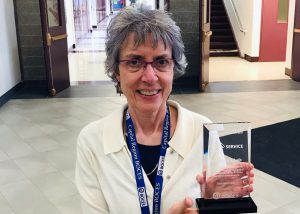 Orientation and Mobility Specialist Katherine Schieb, who has short grey hair and is wearing a white collared cardigan sweater and eyeglasses,looks at and smiles for the camera. Katherine is holding a clear glass award for service to the Capital Region BOCES.