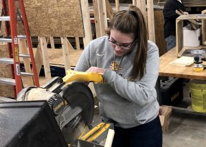 Career & Technical Education Graduate Reagan Smith, who has long, light brown hair pulled back in a ponytail and is wearing eyeglasses and a light grey hoodie, looks down while sawing a piece of wood with a circular saw in a construction and heavy equipment classroom.