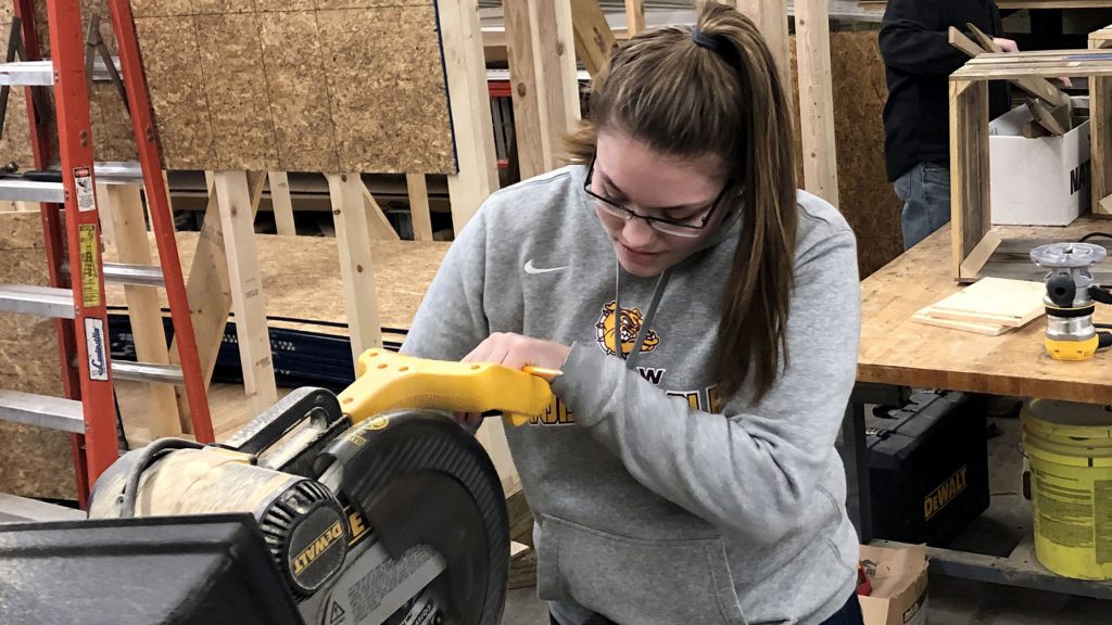 Career & Technical Education Graduate Reagan Smith, who has long, light brown hair pulled back in a ponytail and is wearing eyeglasses and a light grey hoodie, looks down while sawing a piece of wood with a circular saw in a construction and heavy equipment classroom.