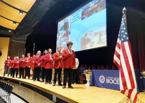 A group of 11 students in red SkillsUSA jackets stand on a stage for the Pledge of Allegiance