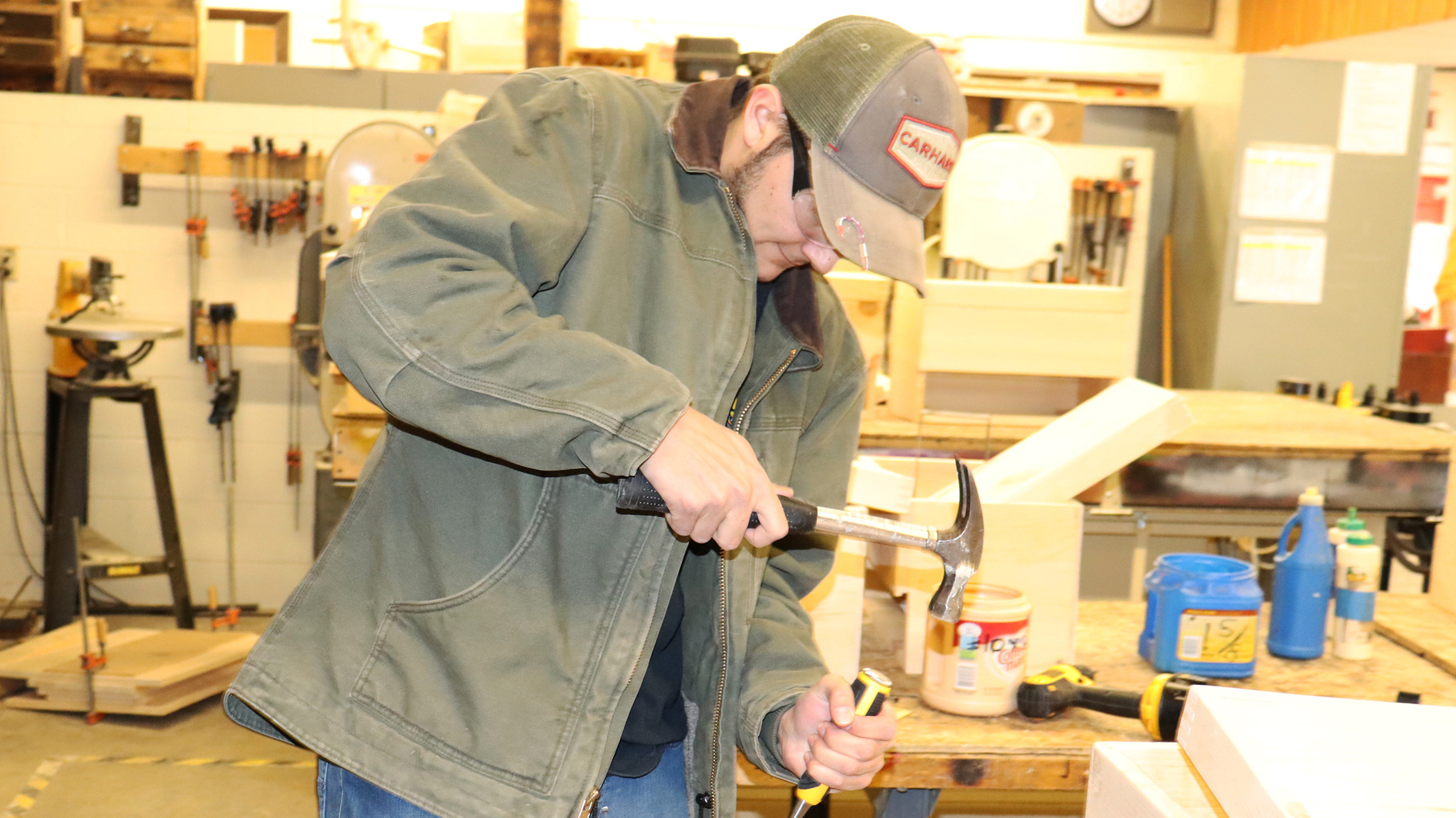 Dillon DiGirolamo, who is wearing a tan Carhartt ball cap and work jacket, blue jeans and protective eyewear, looks downward as using a hammer and chisel to work on a wooden board in a Capital Region BOCES Construction classroom.
