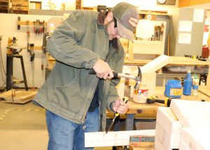 Dillon Digirolamo, who is wearing a tan Carhardtt ball cap and work jacket, blue jeans and protective eyewear, looks downward as using a hammer and chisel to work on a wooden board in a Capital Region BOCES Construction classroom. 