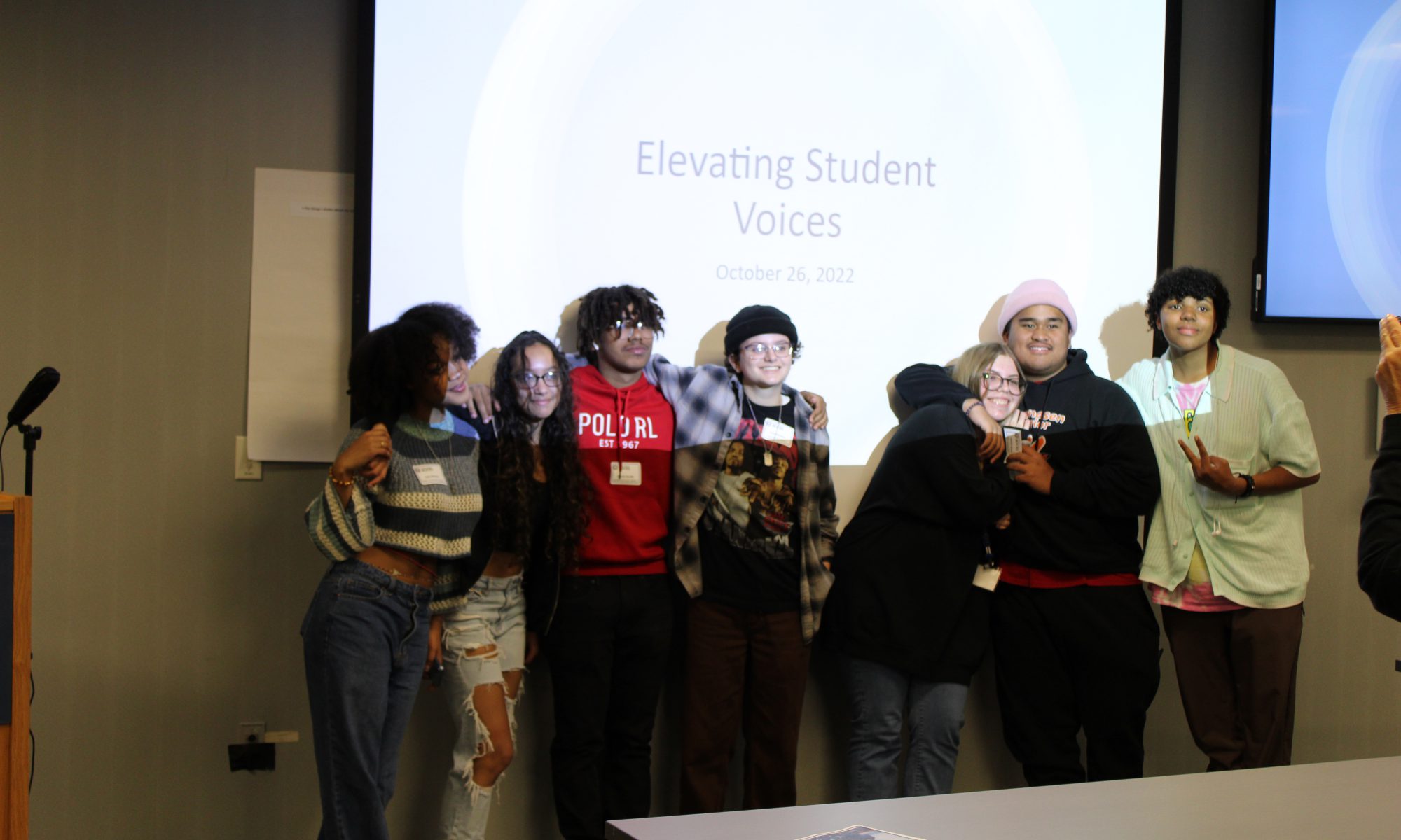Seven students stand in front of projected screen that says "Elevating Student Voices"