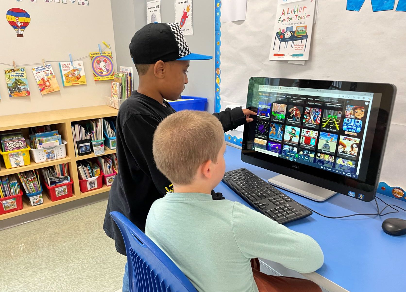 A student wearing a black hat, points to something on a computer screen, while a student in a green shirt sits in front of the screen.