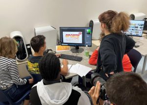 Five Digital Media students looking at a computer screen with an Adobe document displayed that reads "Sports"