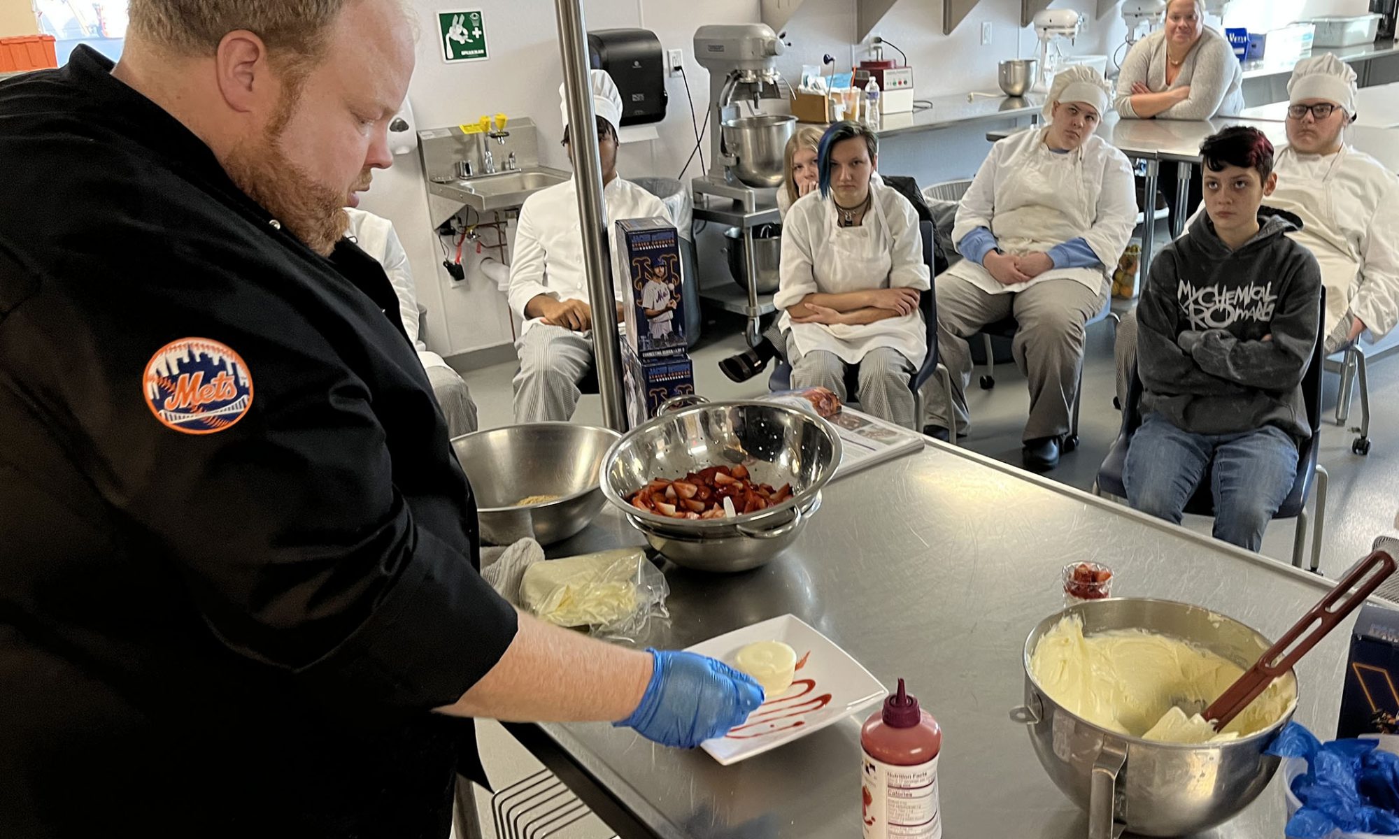 Jason Eksterowicz assembles a strawberry cheesecake, while students are seated and look on. Jason is wearing a black chefs coat with a New York Mets emblem on the sleeve.