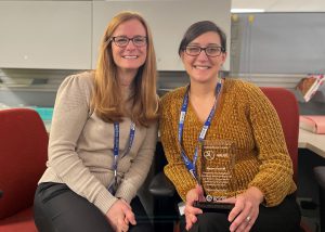 Graphic Designer Karen Pacelli, who has black hair pulled back from her face, is wearing eyeglasses and a mustard colored sweater, and is holding a clear commitment award, sits to the right of program manager Erin McNulty. Both are looking at and smiling for the camera.