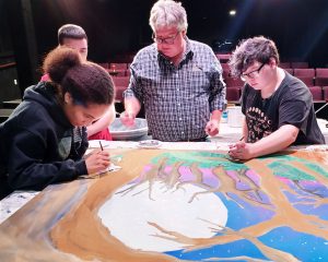 Three students illustrate a forest scene, an instructor watches.