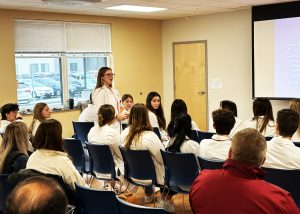 Student Alleda Hawron is wearing a white coat, and is standing speaking to a crowd of seated individuals.