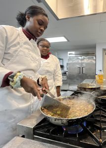 Two culinary students cooking food in a pan. One student holds cooking utensils.