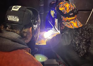 Two individuals wear welding helmets. The individual on the right is welding, and there is a flame.