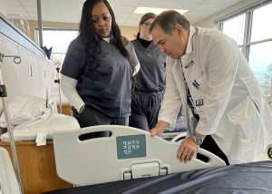 Sharna Smith, an adult LPN student, observes instructor Mark Walh adjust a hospital bed. Smith is wearing grey scrubs. Walh is wearing a white coat and BOCES name tag.