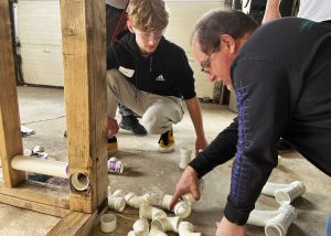 A student and an individual kneeled down, working on pipes.