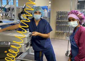 An individual, masked and in scrubs stares at the camera. To the right is another individual masked and in scrubs with a purple jacket. They are in a room at a hospital facility.