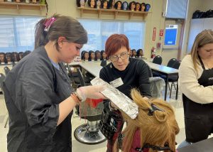 A cosmetology student wearing gloves, holds a foil packet of hair, as an individual, a cosmetology educator, points to a section of hair on a mannequin.