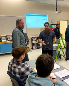 An individual speaks to two seated students, an individual, Jim Haas, observes.