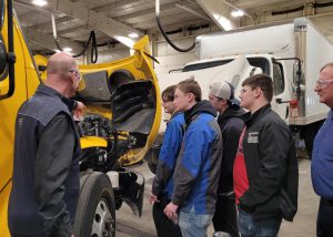 Four students stand next to a yellow Penske truck as an individual points under the hood of the truck.