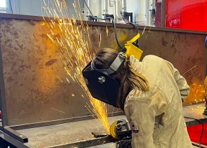 A student in protective clothing and headgear welds. Orange sparks appear.