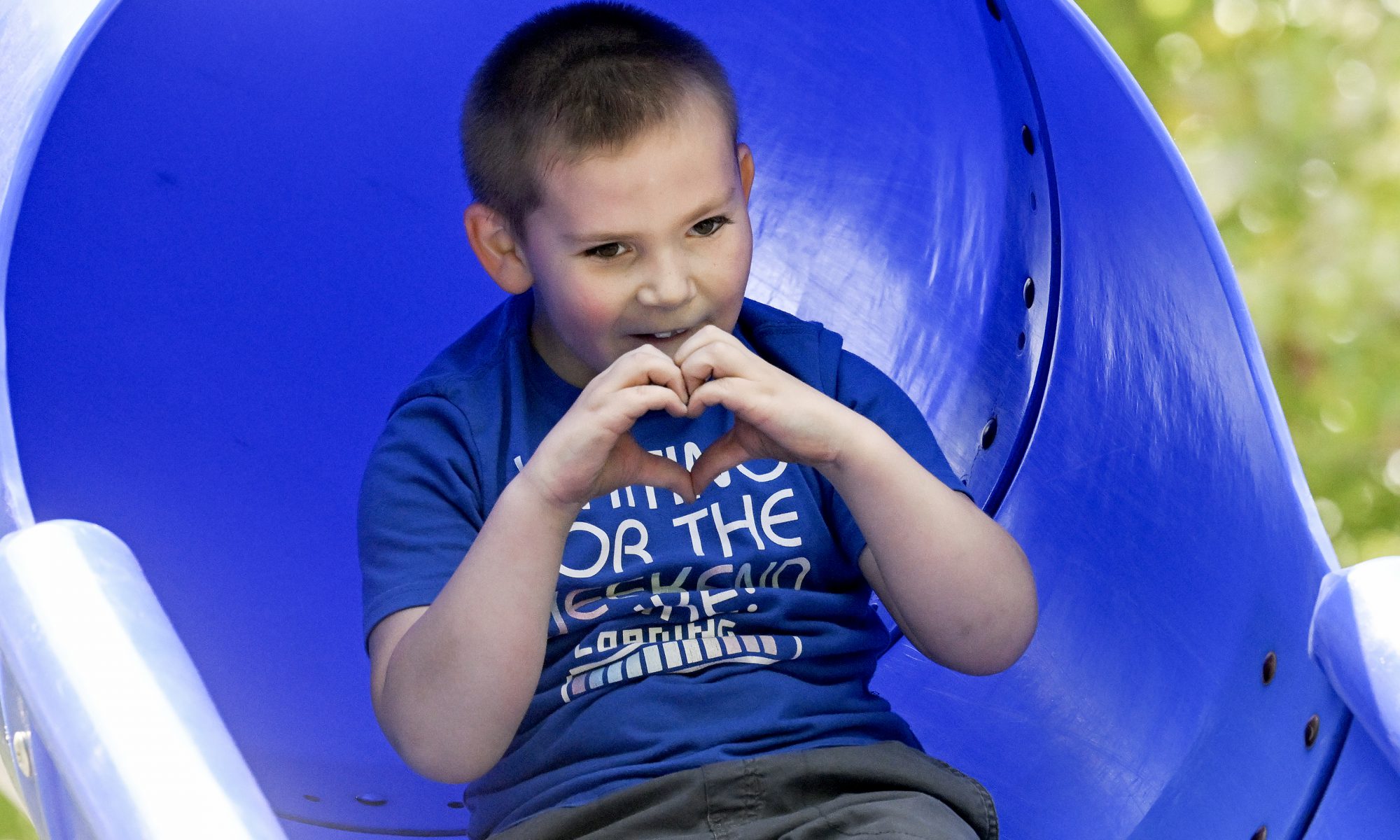 A young childe with close cut brown hair and wearing a bright blue t-shirt pauses at the bottom of a circular bright blue slide. They are smiling and holding their hands together to make the shape of a heart.