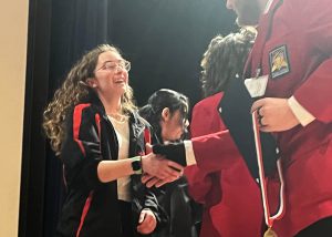 Emily Tice wearing a black and red SkillsUSA jacket shakes the hand of an individual with a red SkillsUSA jacket holding a medal.