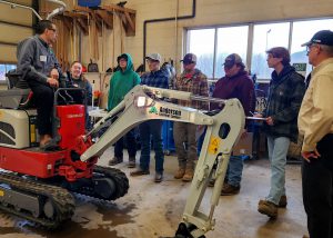 An individual on an equipment vehicle while 7 individuals, 6 students and one teacher, observe.