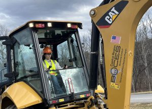 A student in protective vest, hat and goggles operates a yellow heavy equipment vehicle.