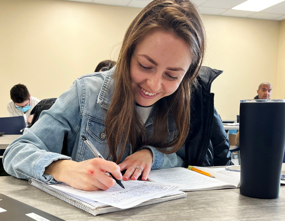 An individual sitting at a table in a classroom with a book open, smiling at the camera.