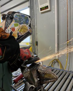 Student Reagan Pedinotti in a protective helmet, welding a metal shark.