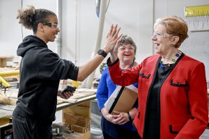 Labor Commissioner Roberta Reardon high-fives student Elijah Edwards. They are standing in a workshop and are both wearing protective goggles.