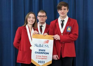 Criminal Justice student Rhiannon Islip stands at left and in a row with two classmates. All are looking at and smiling for the camera. The are wearing bright red blazers, have medals hanging from red white and blue striped ribbons around their necks and are holding a banner that reads "Skills USA State Champion Crime Scene Investigation 2023."