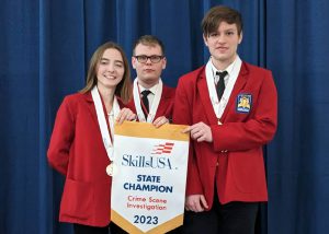 Criminal Justice student Rhiannon Islip stands at left and in a row with two classmates. All are looking at and smiling for the camera. The are wearing bright red blazers, have medals hanging from red white and blue striped ribbons around their necks and are holding a banner that reads "Skills USA State Champion Crime Scene Investigation 2023." 