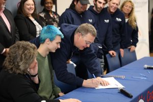 A close up photo of an individual signing a certificate, while nine individuals look on.