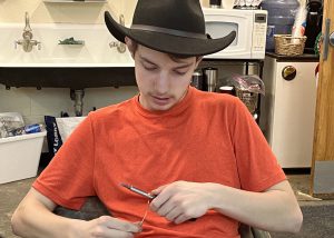 Adult Education students Griffin Boehlke, who is wearing a brown felt cowboy hat and a short-sleeved orange t-shirt, is sitting at a work table, and looking downward while bending wire with a hand tool. 