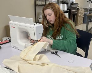 A student sewing a piece of beige fabric using a sewing machine.