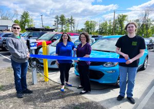 William Kruk and Gavin Christman hold up a blue ribbon while Kate Kruk and Kate Nickerson hold ceremonial scissors. They are standing next to an EV vehicle charging station.