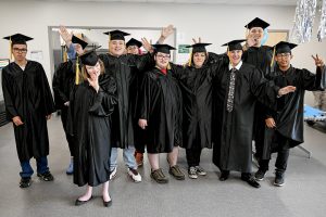 A group photo of 10 students in black graduation regalia smiling and making peace sign gestures.