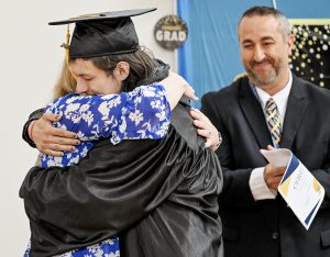 A student in black graduation regalia hugs an individual as principal Gabe Barbato smiles and looks on.